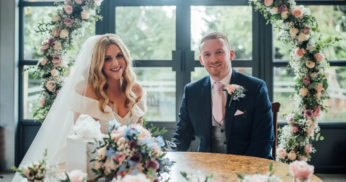 The bride and groom sitting together at their signing table during the ceremony, framed by a semi-circular floral arch. They are both smiling directly at the camera, with a vibrant floral bouquet visible in the foreground.